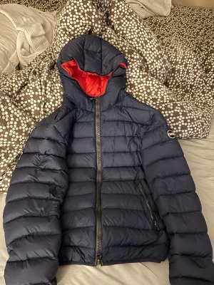 Mörkblå Colmar dunjacka med röd insida perfekt till höst och vår. OBS pris kan diskuteras  - Snygg mörkblå dunjacka från Colmar med röd insida och huva. Jackan har quiltad design, två dragkedjeförsedda fickor och en Colmar-logga på ärmen. Perfekt för kalla vinterdagar och riktigt skön att bära. Materialet är klassiskt dun för maximal värme.