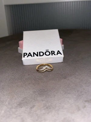 Guldfärgad ring från Pandora - Snygg guldfärgad ring från Pandora med vågig form och glittrande stenar längs hela ovansidan. Ringen har en elegant och modern design som sticker ut och passar perfekt för dig som gillar detaljer. Kommer i originalask från Pandora. Skickas i litet packet/200g