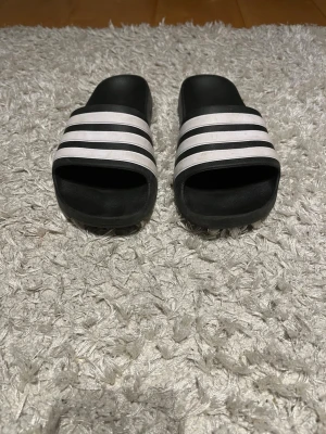 Svarta Adidas sandaler - Säljer ett par klassiska svarta sandaler från Adidas med mönstrad sula och tydlig logga på undersidan. Perfekta att glida runt i och riktigt sköna att ha på sig. Materialet är syntet och designen är enkel och sportig.
