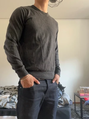 Mörkgrå basic tröja herr - En enkel och stilren mörkgrå tröja med rund halsringning och långa ärmar. Tröjan har ribbade muddar vid ärmslut och nederkant, perfekt för en clean och avslappnad look. Passar till det mesta och är lätt att matcha med andra plagg.