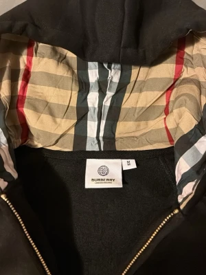 Svart Burberry hoodie med dragkedja - Svart hoodie från Burberry med klassiskt rutigt foder i huvan och guldfärgade detaljer på snören och dragkedja. Modellen har ribbade muddar, fickor framtill och är tillverkad i mjukt material. Perfekt för dig som vill ha en exklusiv och stilren look.