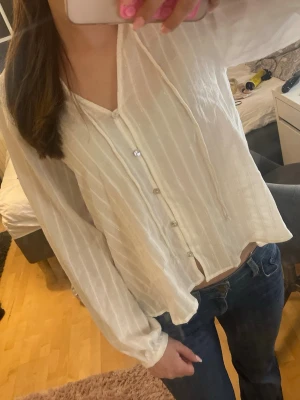Vit blus med knappar och snörning - Säljer en vit blus med subtila ränder, knappar framtill och snörning vid halsen. Blusen har en luftig passform och långa ärmar, perfekt för en avslappnad och trendig look. Materialet är tunt och lätt, vilket gör den skön att bära.