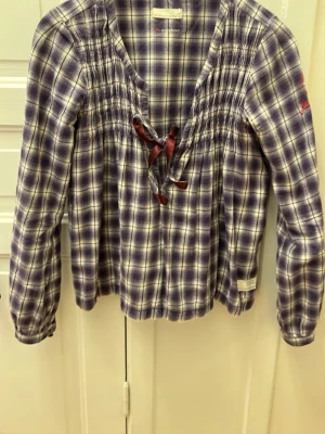 Rutig blus från Odd Molly - Supersnygg rutig blus från Odd Molly i lila och vitt. Blusen har långa ärmar, smock upptill och en dekorativ rosett vid halsringningen. Mjukt bomullstyg som känns skönt mot huden. Perfekt för dig som gillar en avslappnad men trendig stil.