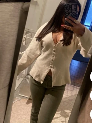 Beige kofta med knappar - Trendig beige kofta med v-ringning och dekorativa knappar framtill. Modellen har långa ärmar och lätt utsvängd passform. Perfekt att styla med jeans eller kjol för en avslappnad look.