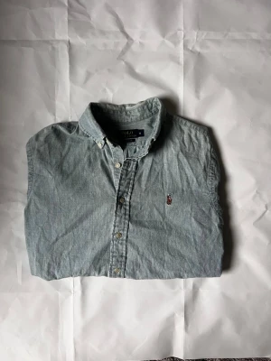 Ljusblå jeans  skjorta från Polo Ralph Lauren - Klassisk ljusblå skjorta från Polo Ralph Lauren med button-down krage och broderad logga på bröstet. Skjortan har långa ärmar och knäppning framtill. Perfekt för en avslappnad men ändå stilren look.
