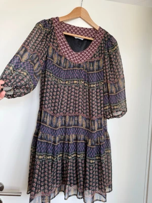 Mönstrad kortklänning med volang - Säljer en luftig kortklänning med blommigt och paisley-inspirerat mönster i flera färger. Klänningen har trekvartsärmar i transparent material, volang nertill och en bred mönstrad kant vid halsen. Perfekt för dig som gillar boho-stil och färgglada detaljer.