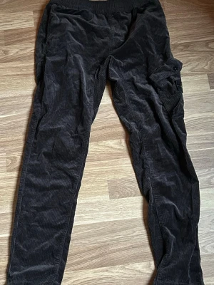 Svarta manchesterbyxor Stone Island - Svarta manchesterbyxor från Stone Island med klassisk patch på benfickan. Byxorna har avsmalnande ben, resår i midjan och vid benslut samt en stor ficka på ena benet. Perfekta för en avslappnad och trendig streetwear-look. Inköpt på nk.se, nypris 2349 kr.