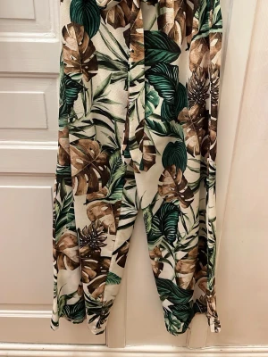 Sommarbyxor med tropiskt mönster - Snygga sommarbyxor med tropiskt blad- och växtmönster i grönt och brunt på ljus botten. Vida ben och avslappnad passform, perfekta för en chill och trendig look. Materialet är linneblandning som känns svalt och skönt. Resår i midjan för extra komfort och skärp. 