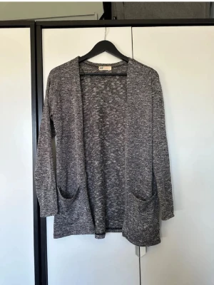 Gråmelerad kofta från H&M - Mysig gråmelerad kofta från H&M med öppen front och två stora fickor framtill. Långärmad och i mjukt stickat material, perfekt att slänga på sig när det är lite kyligt ute.