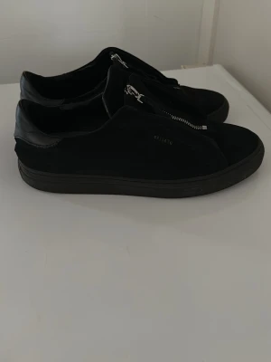 Axel Arigatos  - Stilrena svarta sneakers från Axel Arigato i mocka och läder med coola silverfärgade dragkedjor framtill istället för klassiska snören. Snygg minimalistisk design och låg modell som passar perfekt till streetwear. Väldigt fina och bra skick storlek 42