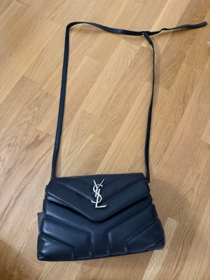 Svart axelväska från Saint Laurent  - Snygg svart axelväska från Saint Laurent med quiltad design och silverfärgad YSL-logga framtill. Väskan har justerbar axelrem och är tillverkad i mjukt läder. Perfekt för dig som vill ha en stilren och lyxig accessoar. Aldrig använd 