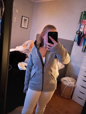 Grå fleecejacka med dragkedja - Mysig grå fleecejacka med hel dragkedja framtill och två snedställda fickor med svarta detaljer. Jackan har huva och en avslappnad passform, perfekt för kyliga dagar. 