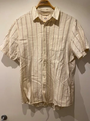 Randig kortärmad skjorta från Marine Layer - Säljer en beige kortärmad skjorta från H&M med tunna vertikala ränder i rött och blått. Storleken står M/L men passar mer som L.