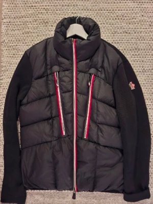 Svart pufferjacka från Moncler Vår/höst  - Cool svart pufferjacka från Moncler med quiltad front och stickade ärmar. Jackan har två vertikala fickor med röda och vita dragkedjor samt Moncler-logga på vänster ärm. Hög krage och snygga detaljer gör den perfekt för kalla dagar.
