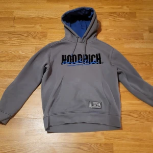 Grå hoodie från Hoodrich - Säljer en grå hoodie från Hoodrich med blått foder i luvan och svart text på bröstet/ryggen.perfekt till både sommar och vinter