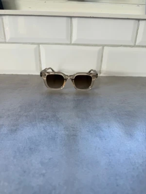Beiga solglasögon från CHIMI 04 - Snygga solglasögon från CHIMI med transparent beige båge och fyrkantig form. Glasen är mörka och ger en cool kontrast till den ljusa ramen. Kommer med originaletui och förpackning. Perfekta för att lyfta din stil i solen. Och dom är som ny men väljer att sälja dom för har inget behov av dom 