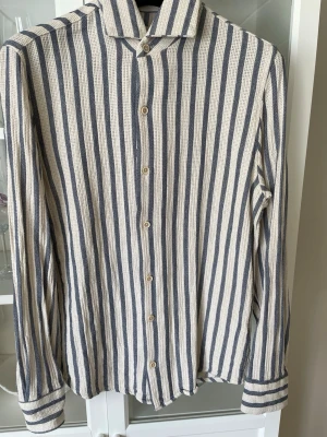 Randig skjorta från Antioch i bomull - Snygg långärmad skjorta från Antioch i 100% bomull. Skjortan har vertikala blå och beige ränder, klassisk krage och knäppning framtill. Perfekt för dig som gillar en avslappnad men stilren look. Ny pris 899kr, Mitt pris: 449kr. Endast använd 2 gånger 