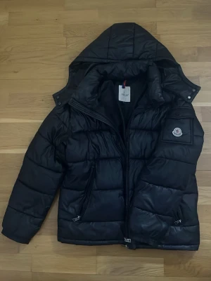 Svart dunjacka från Moncler - Snygg svart dunjacka från Moncler med klassisk logga på ärmen. Jackan har huva, två dragkedjefickor och quiltad design. Det är bara att höra av sig om ni undrar något och på bilden kan man se det som är tyvvär lite slitet på jackan ( knapparna på armen och häng-saken i nacken) så det är därför jag säljer den lite billigare, men annars utöver det så är den i bra skick!❤️ Jag tvättar jackan innan den skickas så hör av er🫶🏾