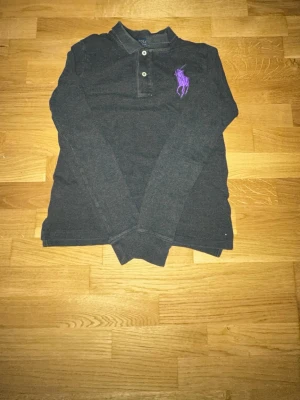 Mörkgrå långärmad henley från Polo Ralph Lauren - Säljer en mörkgrå långärmad henley från Polo Ralph Lauren med lila broderad logga på bröstet. Skjortan har knappar vid halsen och är tillverkad i mjuk bomull. Perfekt för en avslappnad men ändå snygg stil.