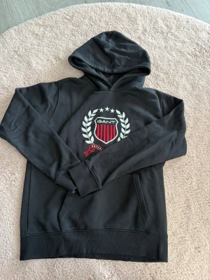 Svart hoodie från Gant med emblem - Snygg svart hoodie från Gant med stort broderat emblem på bröstet i vitt och rött. Hoodien har huva, ribbade muddar och magficka. Perfekt för dig som gillar klassisk college-stil och vill ha något bekvämt och stilrent.
