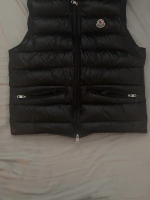 Svart dunväst från Moncler - Snygg svart dunväst från Moncler med glansig finish och klassisk logotyp på bröstet. Västen har två dragkedjeförsedda fickor och en robust dragkedja framtill. Perfekt för lager-på-lager och streetstyle.