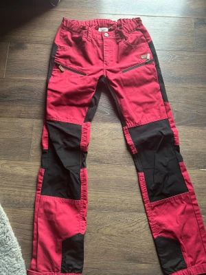 Rosa och svarta friluftsbyxor - Snygga röda friluftsbyxor med svarta förstärkningar på knän och ben, samt dragkedjefickor framtill. Byxorna har elastisk midja och är perfekta för utomhusaktiviteter. Materialet är slitstarkt och passar dig som gillar äventyr. I storlek 159-164