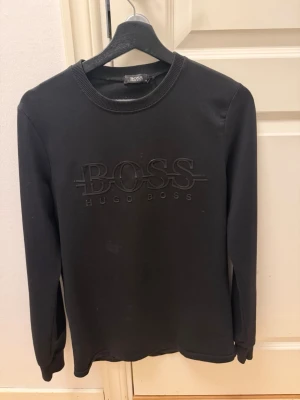 Svart sweatshirt från Hugo Boss - Svart sweatshirt från Hugo Boss med stort BOSS-tryck på bröstet. Tröjan har rund halsringning och långa ärmar. Tillverkad i mjukt material som känns skönt mot huden. Perfekt för en stilren och avslappnad look.