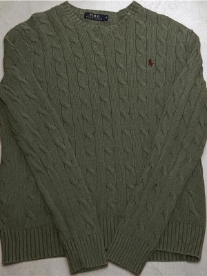 Grön kabelstickad tröja Polo Ralph Lauren - Kabelstickad tröja i olivgrönt från Polo Ralph Lauren. Klassisk rund halsringning, ribbade muddar och diskret broderad logga på bröstet. Perfekt för dig som gillar stilrena och tidlösa plagg med snygg struktur.