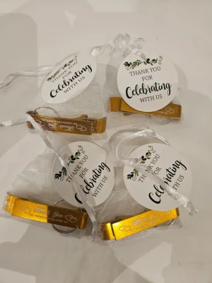 Guldfärgade nyckelringar med tacketikett - Fyra guldfärgade nyckelringar i metall med texten 'Thank You for celebrating with us' och hjärtmotiv. Varje nyckelring ligger i en vit organzapåse med rund etikett och dekorativt band. Perfekt som gåva vid fest eller bröllop.