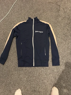 Mörkblå zip-hoodie från Palm Angels - Snygg mörkblå hoodie från Palm Angels med dragkedja framtill och beigea ränder längs ärmarna. Tröjan har hög krage, två fickor med dragkedja och vit logga på bröstet. Perfekt för en streetwear-look.