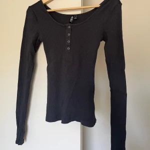 Grå ribbad topp - Svart långärmad topp från Asos med ribbad struktur och rund halsringning. Toppen har knappdetaljer framtill och är figurnära i modellen. Perfekt att styla med jeans eller kjol för en enkel och snygg look. Använd en gång. 