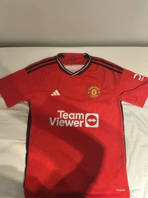 Manchester United matchtröja Adidas - Röd Manchester United matchtröja från Adidas med klubbmärke på bröstet, svarta detaljer och TeamViewer-logga på framsidan. Tröjan har korta ärmar, svart krage och är tillverkad i lätt funktionsmaterial. Perfekt för fotbollsträning eller att bära på matchdag.