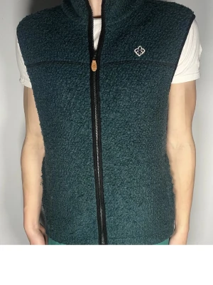 Morris väst olive grön  - Mysig mörkgrön fleeceväst med hel dragkedja framtill och hög krage. Västen har ett broderat vitt emblem på bröstet och är tillverkad i ett fluffigt, teddy-liknande material. Perfekt att slänga över en t-shirt för extra värme.