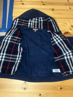 Marinblå hoodie från Burberry - Snygg marinblå hoodie från Burberry med dragkedja och klassiskt rutigt foder i huvan. Hoodien har två fickor framtill, ribbade muddar och justerbar dragsko i huvan. Perfekt för en stilren och avslappnad look. Storlek XL men passar mer som L