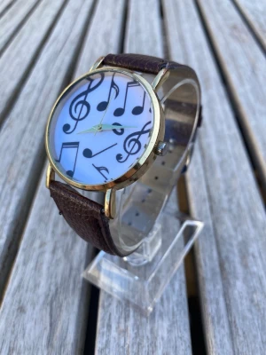 The Music Note Gold 38mm Damur - Armbandsur med ett unikt urtavla prydd med musiknoter. Klockan har ett brunt läderarmband och ett guldfärgat boett. Urverket är kvarts. Mycket gott skick enligt bilder. Behöver batteri.  Nypris: 500 SEK Mitt pris: 189 SEK  Skickas alltid snabbt och spårbart. Skriv vid frågor och se gärna mina andra annonser!