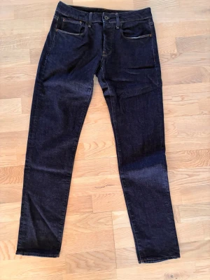 G-Star Mörkblå slim fit jeans med knappgylf - Säljer ett par mörkblå slim fit jeans med knappgylf och klassiska fem fickor. Jeansen har kontrastsömmar i orange och är tillverkade i ett robust denimtyg. Perfekta för dig som gillar en clean och tidlös look.