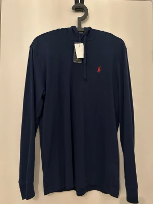 Polo Ralph Lauren  - Mörkblå tröja med ett röt polo Ralph Lauren märke på bröstet. Den har en luva som går att dra åt med hjälp av snörena.