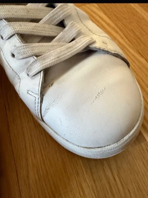 Vita låga sneakers i läder - Snygga vita sneakers i läder med klassisk design och vita skosnören. Skon har en låg profil och svart detalj vid hälen. Perfekt för en clean och stilren look i storlek 41. Skorna har slitage, se bilder. Skickar med hälskydd, se bild. Pris kan diskuteras 