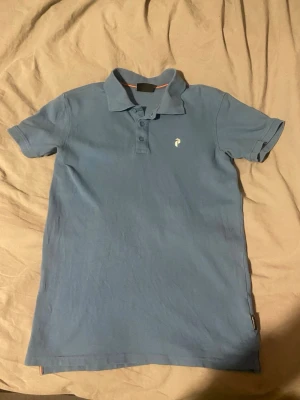 Peak Performance  - Snygg blå pikétröja med klassisk krage och tre knappar framtill. Tröjan har korta ärmar och en liten vit broderad logga på bröstet. Perfekt för en avslappnad men ändå stilren look.