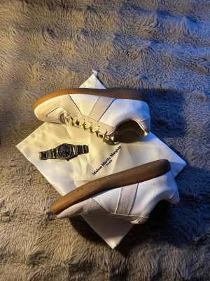 Maison Margiela vita sneakers - Maison Margiela sneakers i vitt läder med beige mockadetaljer och klassisk gummisula. Lågt skaft och stilren design med vita snören. Perfekta för dig som gillar minimalistisk och trendig look.