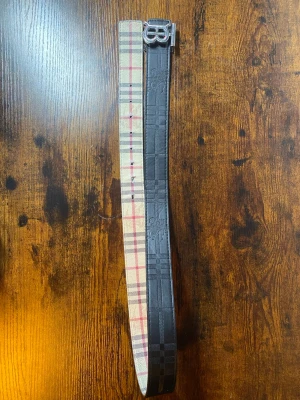 Burberry vändbart bälte med B-spänne - Snyggt vändbart bälte från Burberry med klassiskt rutigt mönster på ena sidan i beige, rött och svart samt svart mönster på andra sidan. Stort B-format metallspänne framtill. Perfekt accessoar för att lyfta din outfit.        ‼️OBS: 3 sista hålet är lite slitna samt lite slitet på B spännet‼️