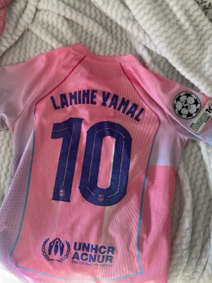Rosa FC Barcelona matchtröja Nike - Säljer en rosa FC Barcelona matchtröja med Lamine Yamal och nummer 10 på ryggen. Tröjan har tryckta loggor från Nike och Barcelona, samt UNHCR och Spotify som sponsorer. Materialet är lätt och andas, perfekt för fotboll eller träning.