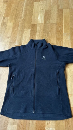 Svart fleecejacka från Haglöfs - Mysig svart fleecejacka från Haglöfs med hel dragkedja framtill och två sidofickor med dragkedja. Jackan har hög krage och diskret Haglöfs-logga på bröstet. Perfekt för lager-på-lager och outdoor-äventyr.