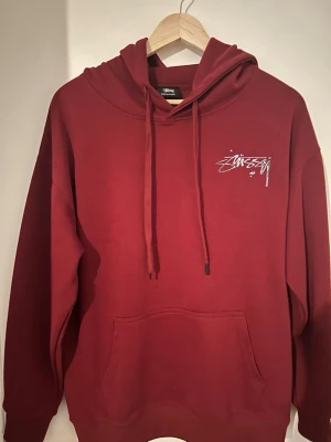 Stüssy hoodie  - Snygg vinröd hoodie från Stüssy med vit logga på bröstet och stor logga i silver på ryggen. Klassisk känguruficka framtill, justerbar huva med dragsko och mjukt material. Perfekt för en avslappnad streetwear-look.