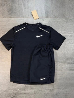 Svart träningsset från Nike - Snyggt svart träningsset från Nike med t-shirt och shorts. Båda plaggen har den klassiska vita Nike-loggan och t-shirten har vita detaljer vid axlarna. Perfekt för träning eller sportaktiviteter. Materialet är lätt och andas bra.
