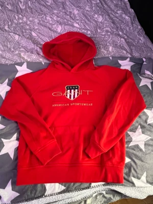 Röd hoodie från GANT med tryck - Snygg röd hoodie från GANT med klassiskt tryck och logga på bröstet. Hoodien har huva, magficka och ribbade muddar. Perfekt för dig som gillar sportig och avslappnad stil. Tillverkad i mjukt material för extra komfort.