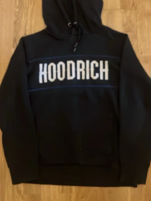 Svart hoodie från Hoodrich - Säljer en svart hoodie från Hoodrich med stor vit logga på både fram- och baksidan. Hoodien har huva med dragsko och en klassisk känguruficka framtill. Materialet känns mjukt och bekvämt, perfekt för en chill och street-inspirerad stil.