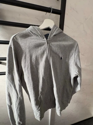 Grå hoodie från Polo Ralph Lauren - Snygg grå hoodie från Polo Ralph Lauren med dragkedja framtill och klassisk huva. Tröjan har två fickor och den ikoniska lilla loggan broderad på bröstet. Tillverkad i mjukt bomullsmaterial som är skönt att ha på sig. Perfekt för en avslappnad och stilren look.