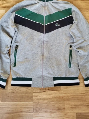 Lacoste kofta - Fin och skön zip-kofta från Lacoste i grått med gröna och mörkblå detaljer. Klassisk och sportig stil som passar bra till vardag och är lätt att matcha med olika outfits.                                                                                             Har dragkedja framtill och två fickor med dragkedja. Mjuk och bekväm att ha på sig. Logga på bröstet.