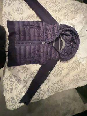 Mörkblå Moncler cardiganjacka. Storlek M - Snygg mörkblå cardiganjacka från Moncler med quiltad front och huva. Jackan har stickade ärmar och sidor, dragkedja framtill och två fickor. Perfekt för lager-på-lager och har en modern, sportig look med Moncler-logga på ärmen. Använd 1 gång, men då jag ändrat min stil vil jag ej ha kvar denna!